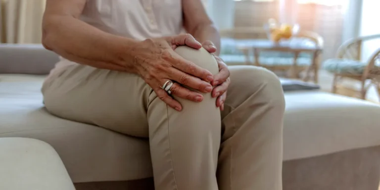 An older adult sitting on a sofa, holding their knee with both hands, suggesting the knee pain of osteoarthritis
